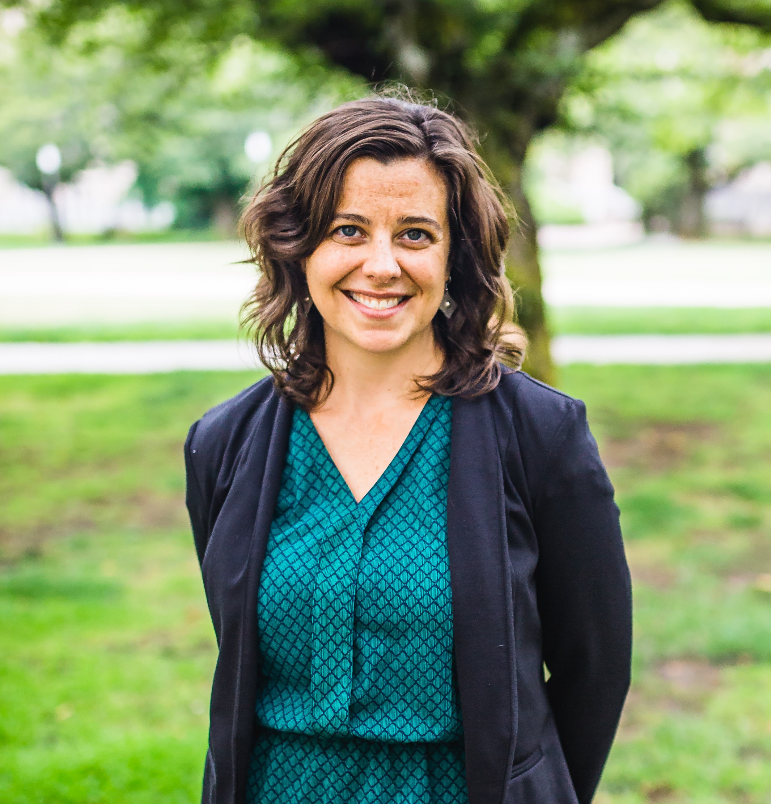 Renee standing and smiling with her hands behind her back. She is wearing a blue diamond patterned dress and a black blazer. She is standing in front of a green field.