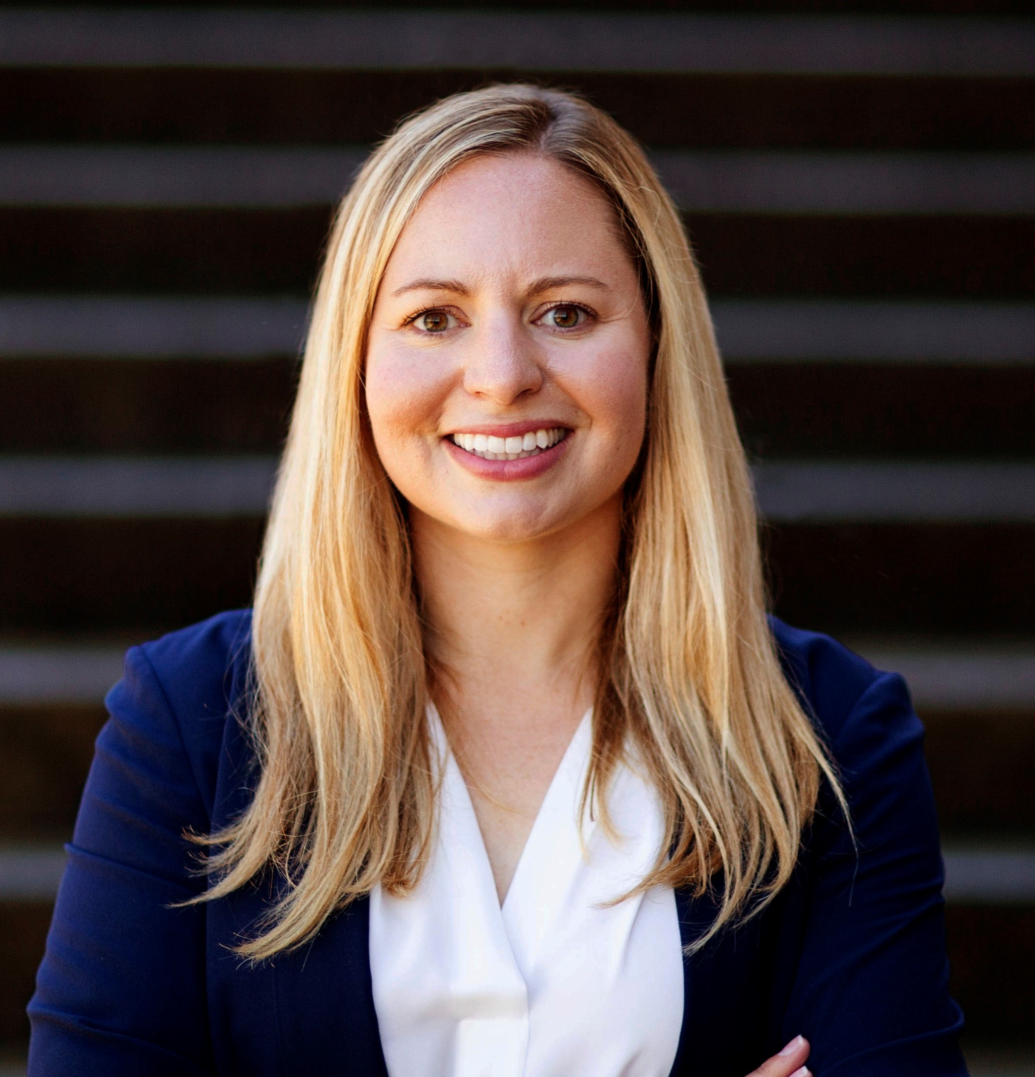 Allison is standing in front of a black and grey striped background. She has her arms crossed and is smiling.