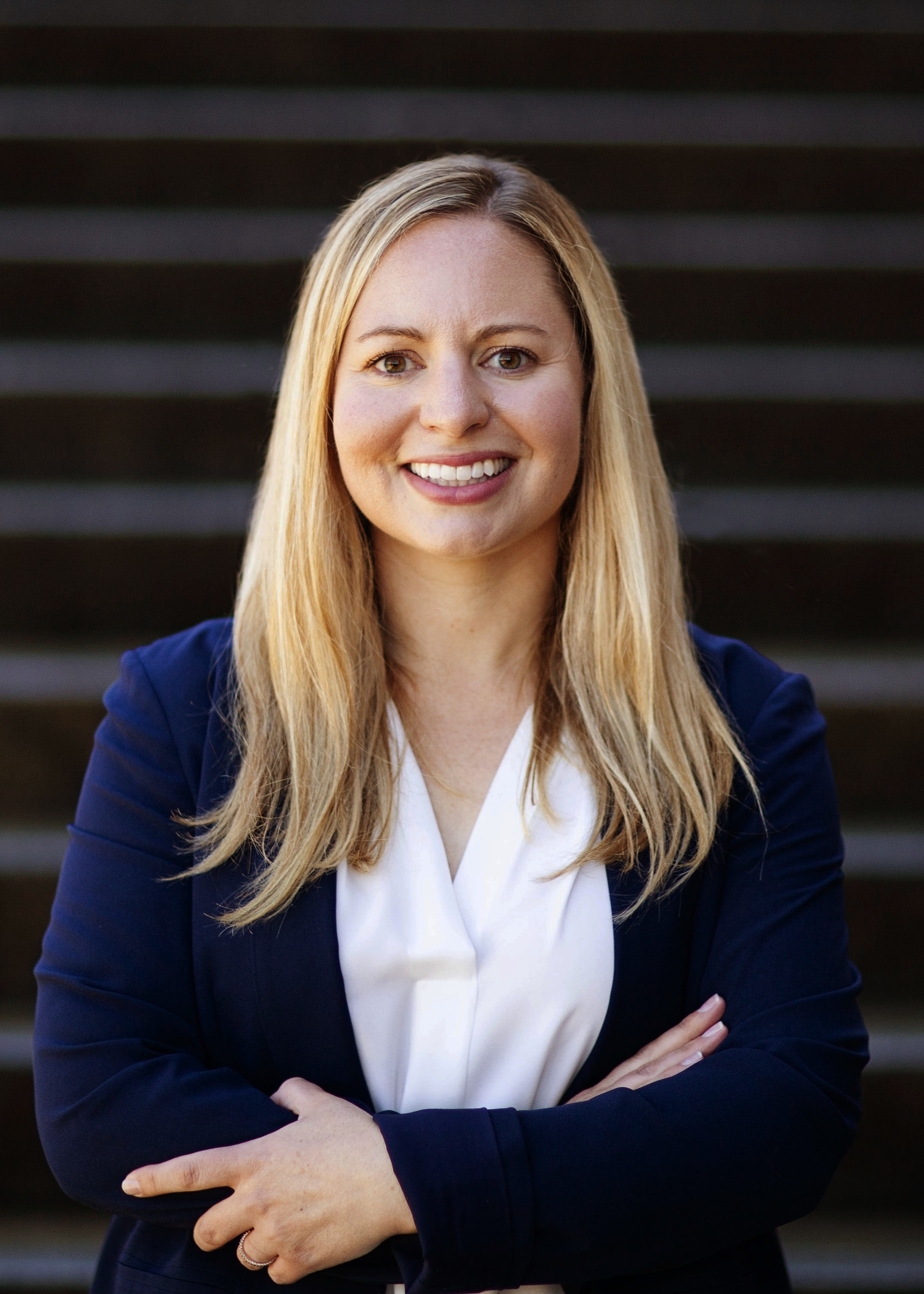 Allison is standing in front of a black and grey striped background. She has her arms crossed and is smiling.