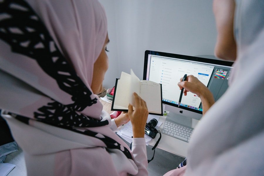 Two women wearing hijabs looking at a computer and a notebook
