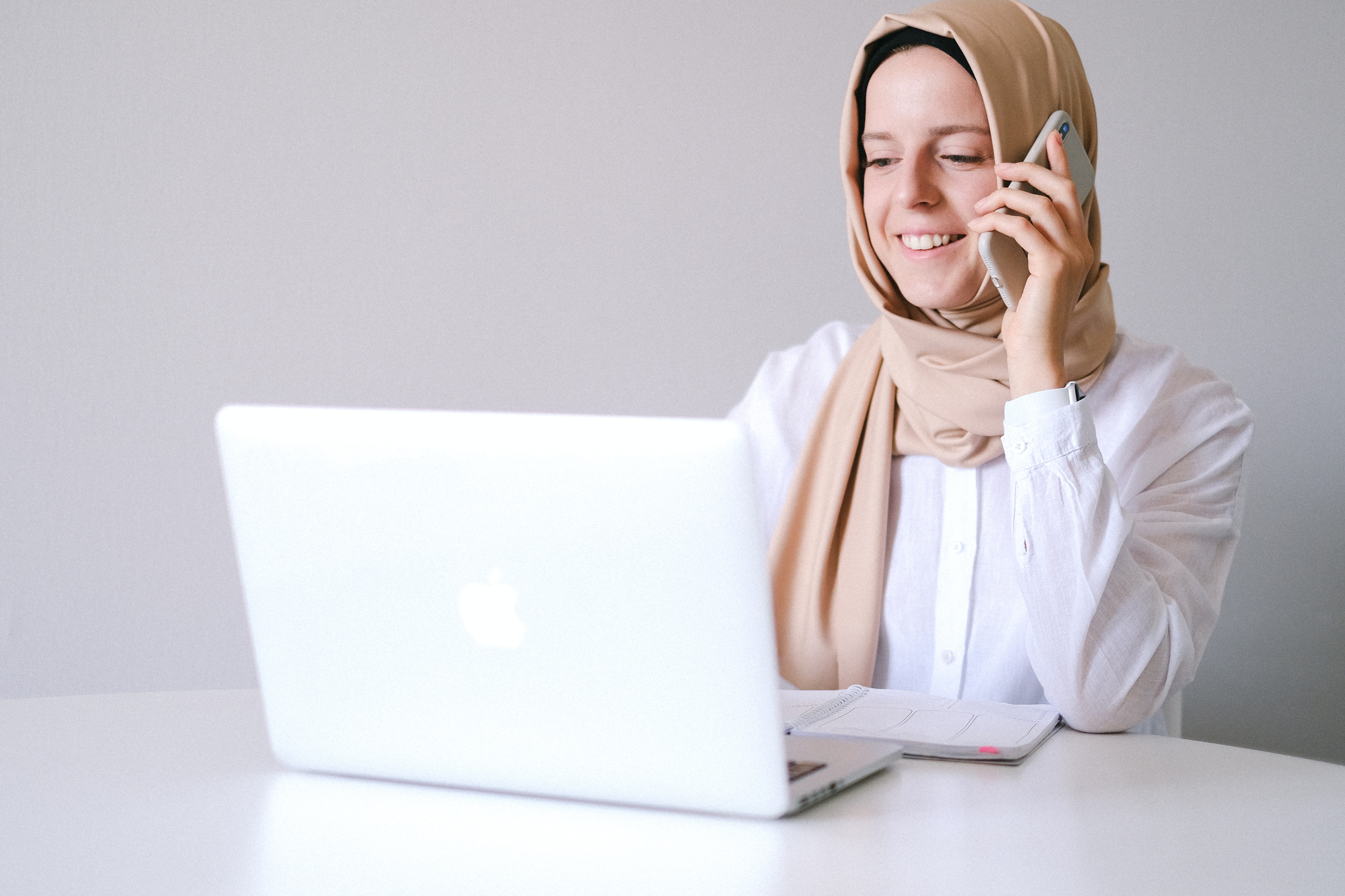 A woman sitting in front of her computer and talking on her mobile phone. She is wearing a white button up shirt and a tan headscarf.