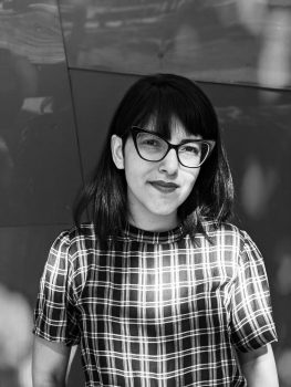 A black and white photo of Itza. She has dark hair with bangs and is wearing glasses and a checkered shirt.