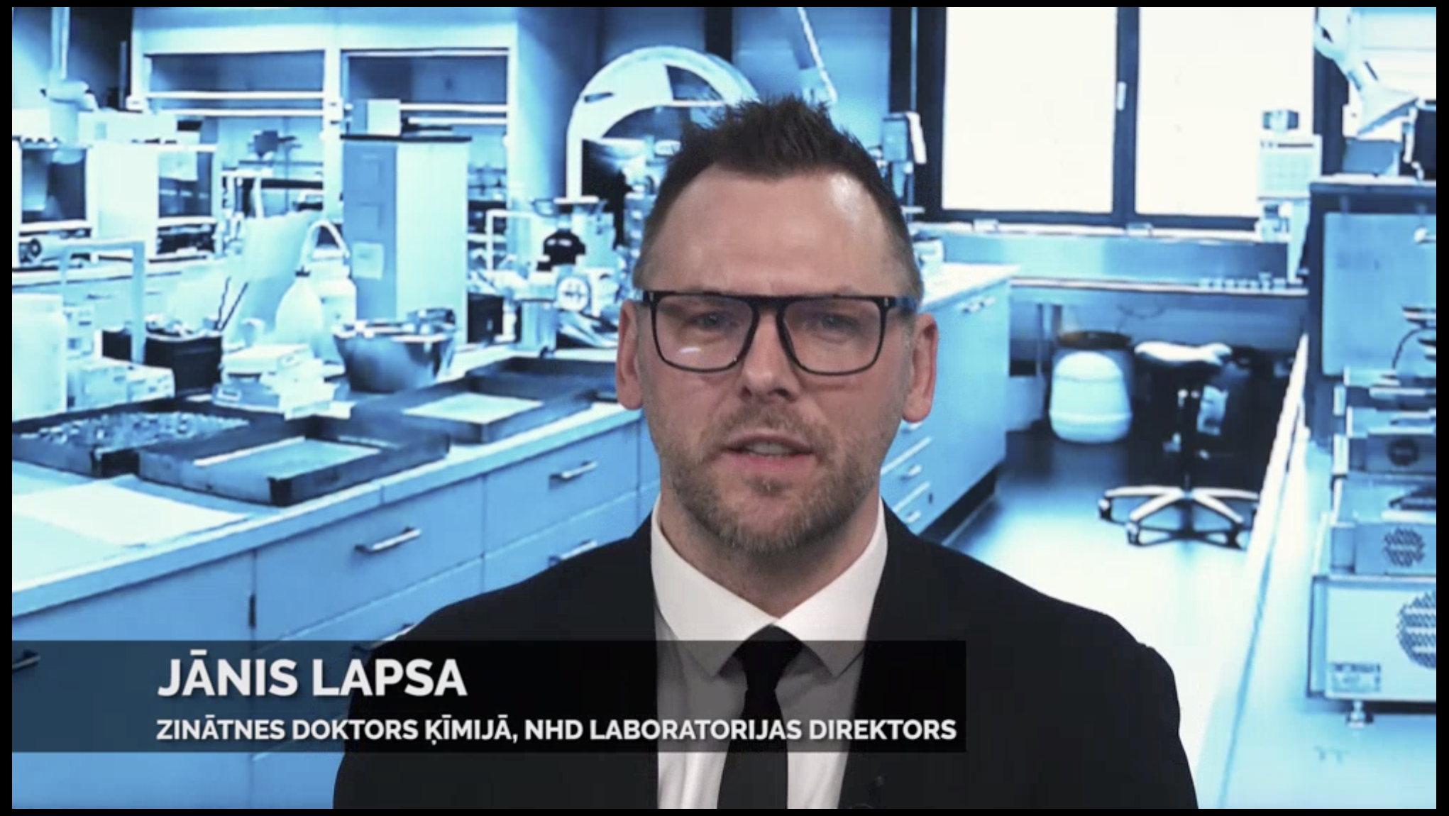 A still frame of a video with a man wearing glasses, a tie, shirt and suit jacket with the title Janis Lapsa Zinatnes doktors kimija, NHD Laboratorijas Direktors in the lower third of the screen