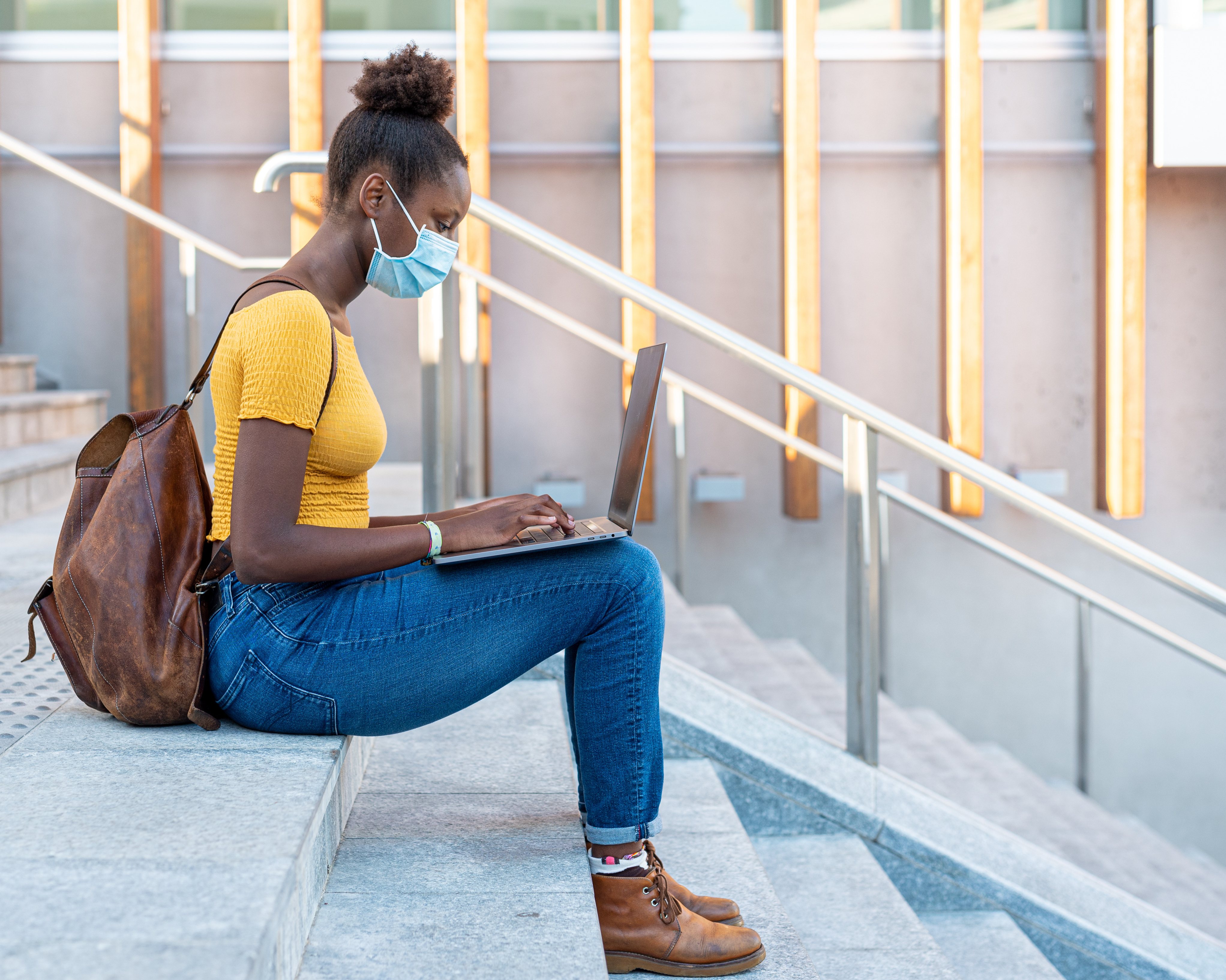 Young person wearing a yellow shirt, jeans, boots, a medical face mask and a backpack sitting on exterior concrete steps typing on a laptop